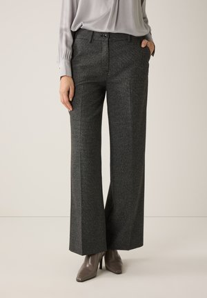 Frau trägt graue Hosen mit Fischgrätmuster und weitem Bein, hellgraue Bluse und taupefarbene, spitz zulaufende Stiefeletten mit Absatz, stehend vor einem einfarbigen Hintergrund.