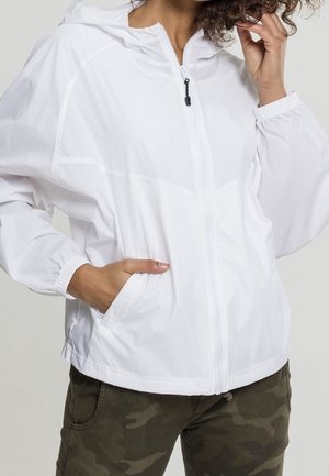 Personne portant une veste blanche à capuche avec fermeture éclair, des poignets élastiques, une main dans la poche, et un pantalon à motif camouflage.
