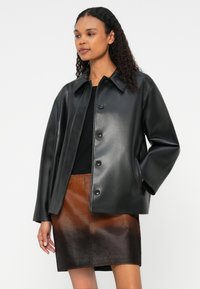 Casaco de imitação de pele preto, com gola, botões frontais e bolsos laterais, combinado com uma saia castanha com textura em degradê.