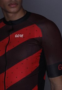 Jersey de ciclismo de manga corta en rojo y negro, con rayas diagonales y lunares, con cremallera completa y el logo de GORE en el pecho.