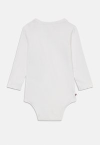 Hosszú ujjú, puha pamutból készült fehér bodysuit, kerek gallérral és patentgombos záródással az alján. Az oldalon a márka színeivel ellátott címke található.
