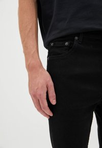 Jeans de mezclilla negra con corte ajustado, que cuentan con un bolsillo delantero, botones de metal y una tela ligeramente texturizada. Mano descansando sobre el muslo.