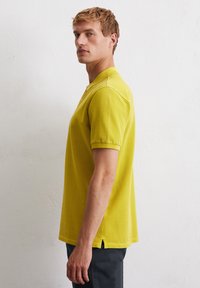 Żółta koszulka polo z klasycznym kołnierzykiem, krótkimi rękawami, bocznymi rozporkami i miękką, teksturowaną tkaniną. Noszona z ciemnymi spodniami.