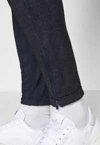 Marinblå rutiga byxor i en texturerad fabrik, med en dragkedjefunktion vid ankeln, kombinerat med vita sneakers och vita strumpor.