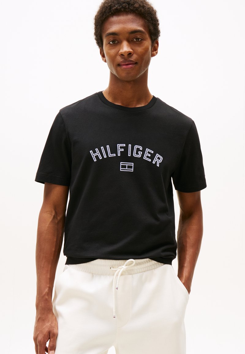 Jeune homme portant un t-shirt noir avec le texte et le logo "HILFIGER", associé à un pantalon blanc à cordon de serrage, se tenant avec une main dans la poche.