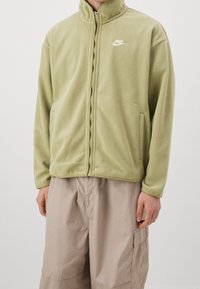 Fleece zipová bunda ve světle olivové barvě s vysokým límcem, dvěma bočními kapsami a malým bílým logem Nike na levé straně hrudi.