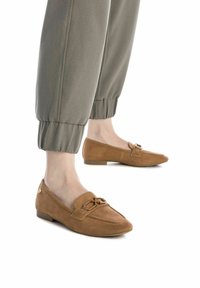 Mocasines de ante marrón con un tacón bajo, que cuentan con una correa decorativa y detalle de hebilla. Tienen una punta afilada y una superficie texturizada.