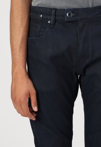 Dark blue denim jeans with a fitted design, featuring a front pocket, metal button, and contrast stitching. Smooth texture with subtle sheen.