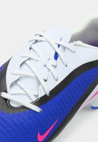 Ποδοσφαιρικό παπούτσι Nike με λευκό επάνω μέρος, μαύρες και μπλε λεπτομέρειες, υφή σχεδίασης, λευκές κορδόνες και ροζ λογότυπο Nike.