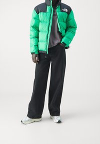 Zelená péřová bunda s černými detaily, s zipem a logem. Nositá přes šedé tričko a spárovaná s širokými černými kalhotami a teniskami.