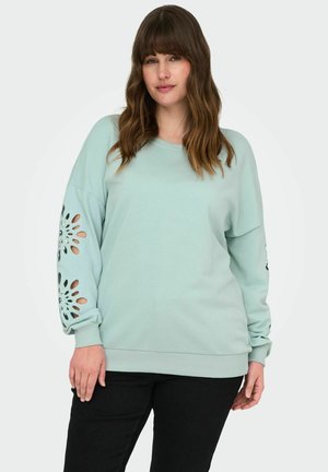 Felpa verde menta chiaro con spalle scese, pannelli frontali e posteriori, e ritagli decorati sulle maniche, abbinata a pantaloni neri.