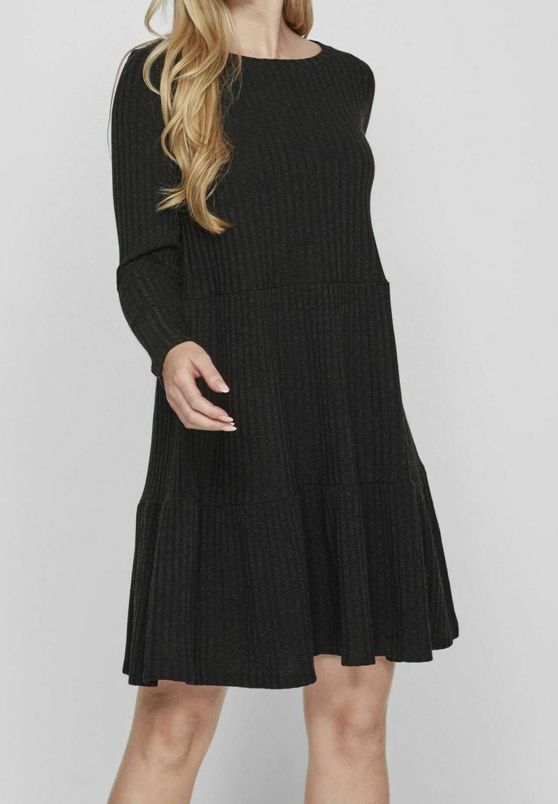 Femme portant une robe en maille côtelée noire, à manches longues, longueur genou, avec une jupe à volants, sur un fond uni.