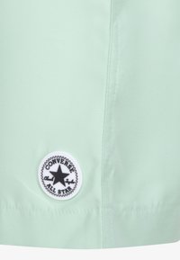 Licht muntgroene stof met een gladde textuur, voorzien van een ronde zwart-witte Converse All Star-patch aan de linksonderkant.