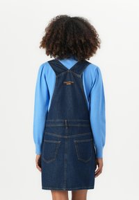 Robe salopette en denim bleu foncé avec bretelles réglables, deux poches avant et une taille ajustée. Présente le logo doré "Marc O'Polo".
