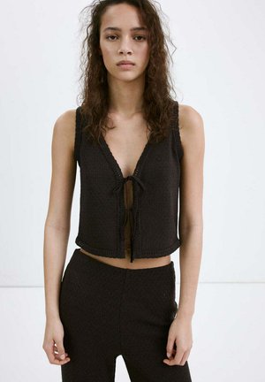 Jeune femme aux cheveux bouclés portant un top noir en maille sans manches noué à l'avant et un pantalon noir assorti sur un fond uni.