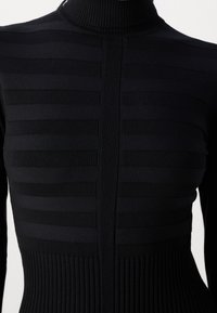 Haut à col roulé noir présentant une texture rayée horizontale sur le haut du corps et un matériau côtelé à la taille. Finition en tissu lisse sur l'ensemble.