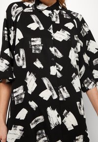 Camicia nera con bottoni e maniche corte, caratterizzata da un motivo astratto a pennellate bianche e un colletto classico. Tessuto di texture liscia.
