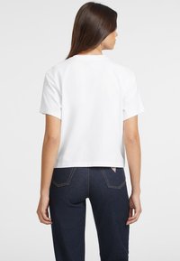 Weißes, kurzes T-Shirt mit rundem Ausschnitt und kurzen Ärmeln, aus einem glatten Stoff gefertigt, kombiniert mit dunkelblauen Jeans mit einem Logofähnchen.