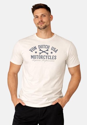 Homme portant un t-shirt blanc Von Dutch USA Motorcycles avec un motif de clés croisées, mains dans les poches de pantalons noirs, fond neutre.