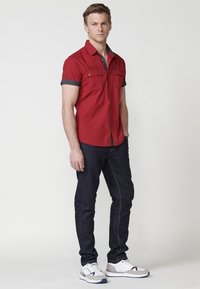 Camisa roja de manga corta con botones y ribete gris, combinada con jeans de mezclilla oscuros y zapatillas deportivas blancas con detalles grises.