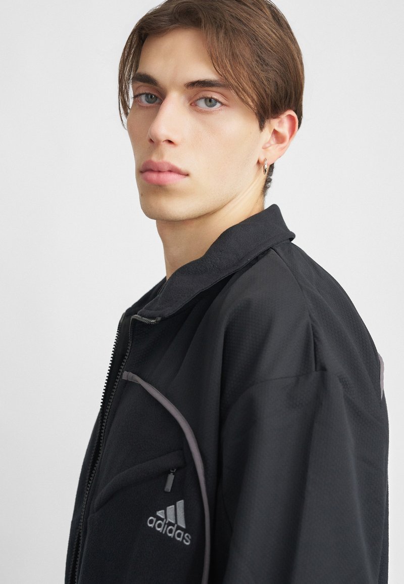 Schwarze Zip-Jacke aus strukturiertem Stoff, grauen Akzenten und einer Vordertasche. Mit einem Adidas-Logo auf der Brust.