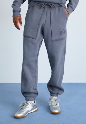 Gray sweatpants with a relaxed fit, featuring two large patch pockets and ribbed cuffs. Paired with silver athletic shoes.