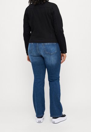 Personne portant une veste noire, un jean bleu et des baskets noir et blanc, debout de dos sur un fond blanc uni.