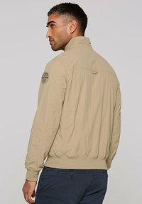 Beige bomber jacket with a high collar, elastic cuffs, and a fitted waistband. Features a patch on the shoulder and a smooth texture.