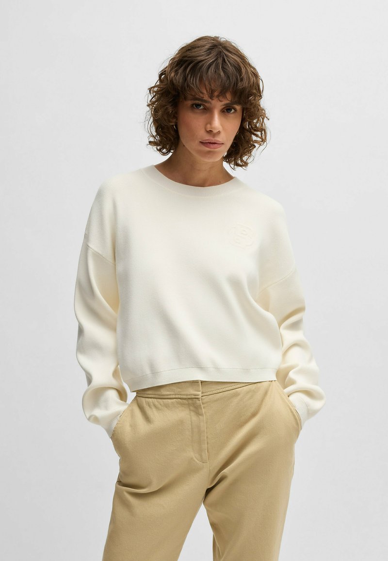 White sweatshirt with a crew neckline, ribbed texture, and embroidered logo on the chest; paired with beige, relaxed-fit pants.