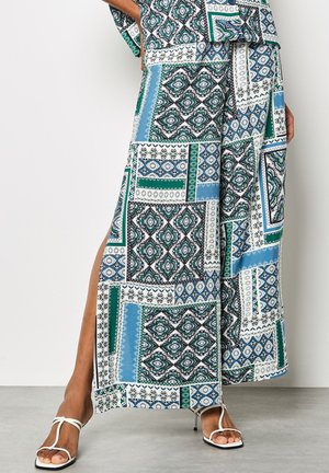 Vrouw draagt wijde broek met patroon en bijpassende top met blauw, groen en zwart geometrisch dessin, gecombineerd met witte sandalen met bandjes.