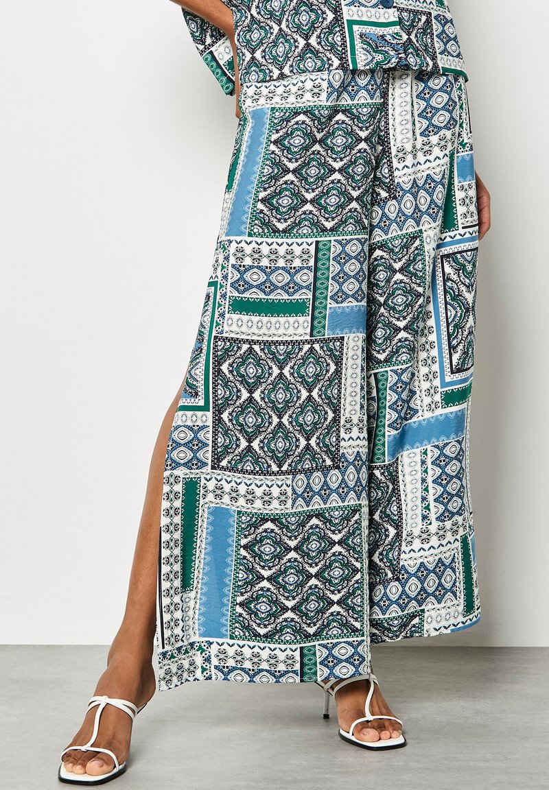 Femme portant un pantalon large à motifs et un haut assorti avec des imprimés géométriques bleus, verts et noirs, associés à des talons blancs à lanières.