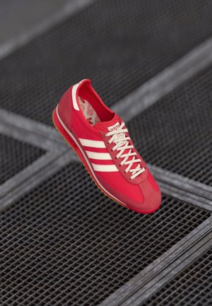 Rode Adidas sneaker met witte strepen en veters, zwevend boven een donker metalen roosteroppervlak.