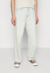 Light blue, loose-fit denim jeans with five pockets, visible stitching, and a frayed edge on the left pocket, paired with brown sneakers.