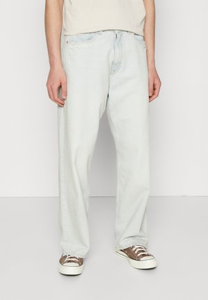 Light blue, loose-fit denim jeans with five pockets, visible stitching, and a frayed edge on the left pocket, paired with brown sneakers.