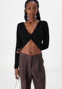 Svart cropped genser med V-hals og vridd frontdetalj, kombinert med høyt liv brune bukser. Glatt tekstur, lange ermer, minimalistisk design.