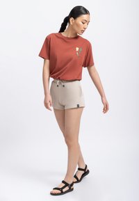 T-shirt rossa in cotone con grafiche verdi e gialle, abbinata a pantaloni beige con coulisse e accenti neri. Sandali neri completano il look.