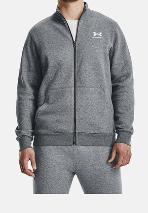 Mężczyzna ubrany w szarą kurtkę zapinaną na zamek marki Under Armour oraz pasujące spodnie, ręce luźno opuszczone wzdłuż ciała, biała koszulka widoczna przy szyi.