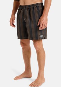 Tmavohnedé plavkové šortky s vertikálnymi prúžkami, elastickým pásom a božím logom. Materiál sa zdá byť ľahký a má hladkú textúru.