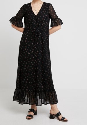 Schwarzes Maxikleid mit kleinem Blumenmuster, transparenten kurzen Rüschenärmeln, Rüschensaum und gebundener Taille, getragen mit schwarzen Sandalen mit Blockabsatz.
