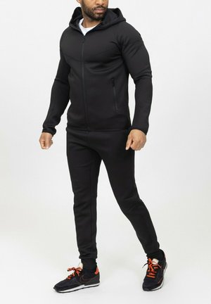 Survêtement de sport noir avec capuche, arborant des poches zippées et une coupe ajustée. Porté avec des chaussures de sport noires et orange. Texture lisse.