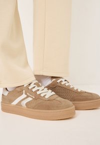 Sneaker in suede marrone con accenti bianchi, pannelli laterali texture e lacci piatti, abbinati a pantaloni chiari e calzini bianchi.