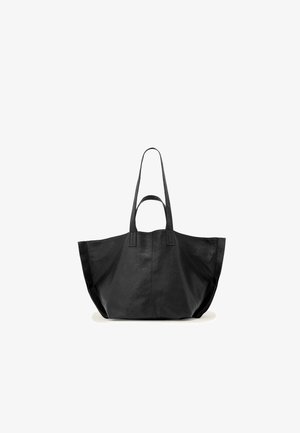 Zwarte leren tote tas met twee lange handvatten en een brede, zachte rechthoekige vorm op een witte achtergrond.