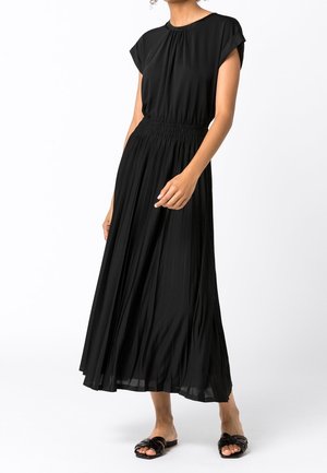 Vrouw draagt een zwarte, geplooide midi-jurk met korte mouwen en een geplooide taille, gecombineerd met zwarte geweven slip-on sandalen.