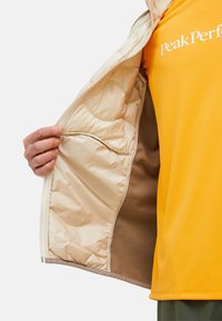 Interno della giacca beige isolata con texture trapuntata; presenta un rivestimento in pile marrone e una tasca laterale. Camicia gialla con logo "Peak Performance" all'interno.