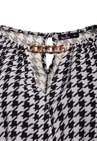 Bluzka w czarno-białą kratę houndstooth z dekoltem w serek i złotym łańcuchem przy szyi. Lekki materiał z marszczonymi detalami.