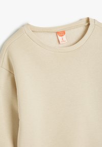 Sudadera beige para bebé con cuello redondo acanalado y textura suave. Presenta una etiqueta naranja con detalles de tamaño. Diseño clásico y sin costuras.