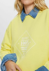 Žlutá mikina z organické bavlny s bílým diamantovým logem a límečkem z denimu. Jemná textura a pohodlný střih.