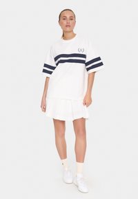 Camiseta blanca de gran tamaño con diseño de rayas navy en el pecho, combinada con una falda blanca plisada y zapatillas blancas. Textura de tela suave.
