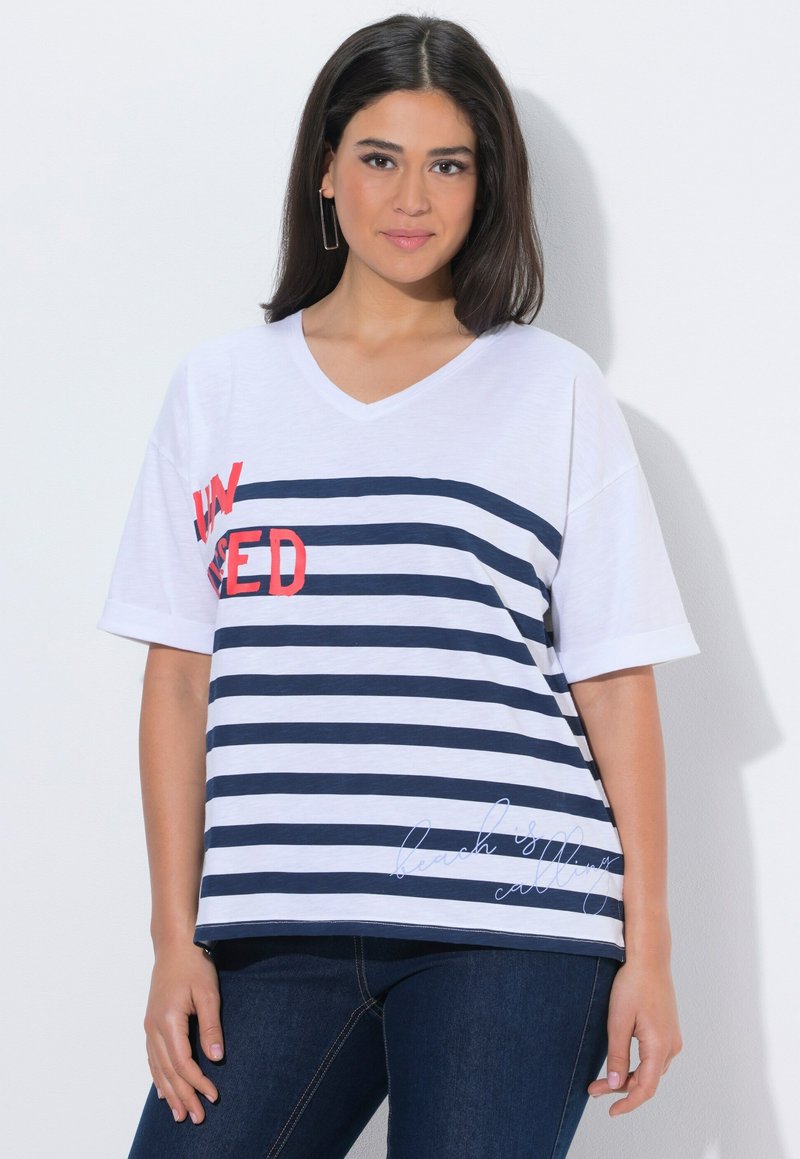 Jeune femme aux cheveux bruns et raides portant un t-shirt à col en V blanc et bleu marine à rayures avec un texte rouge et blanc, associé à un jean bleu foncé.
