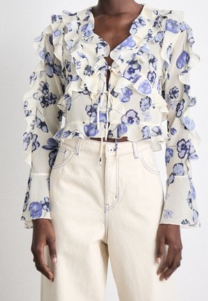 Crèmekleurige blouse met blauw bloemenpatroon, gerimpelde rand, lange pofmouwen en strikjes aan de voorkant, gecombineerd met een hooggetailleerde gebroken witte broek.